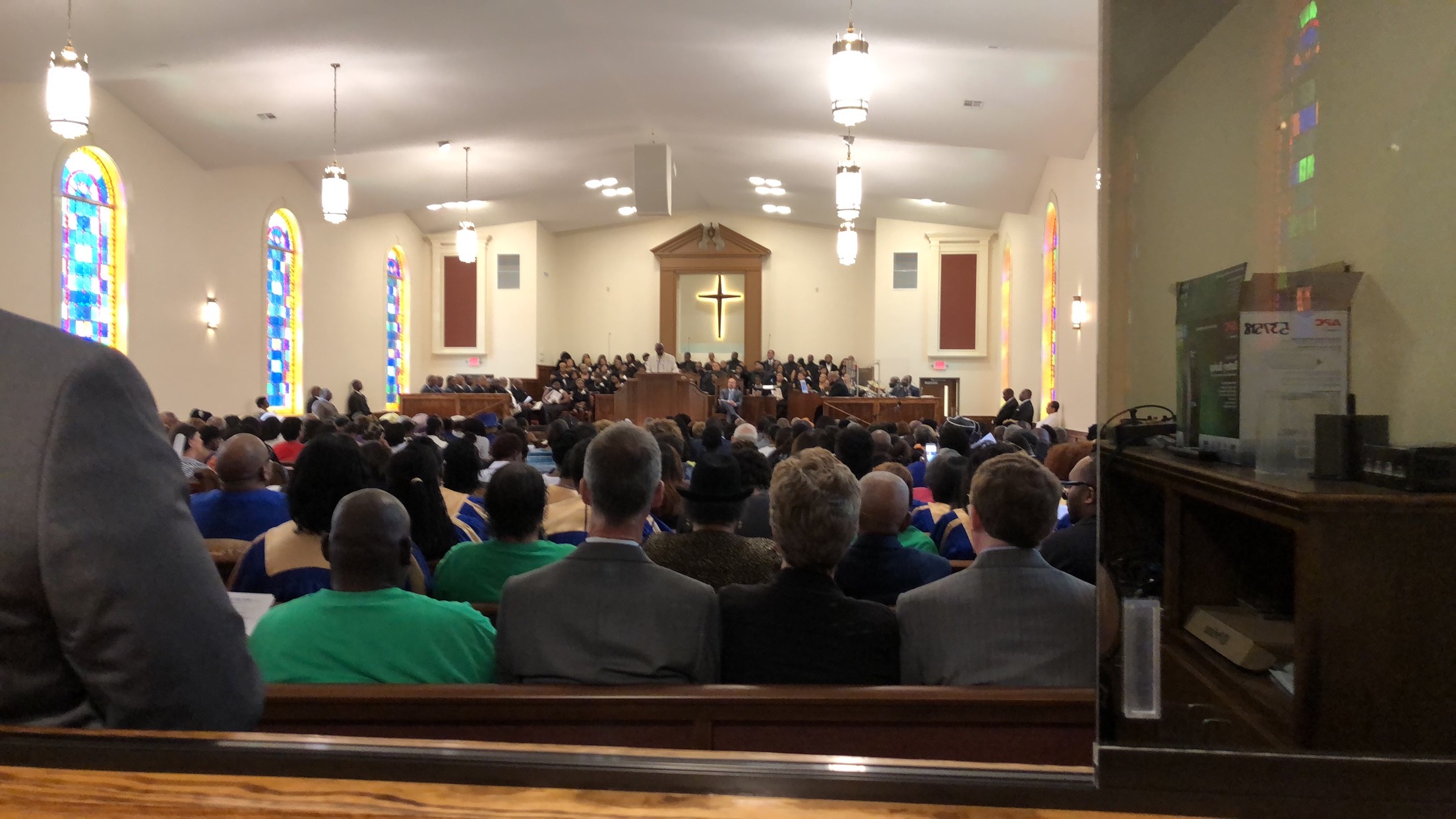 Soundcheck moment with musicians and choir on the platform under the lit cross, white loudspeakers visible hanging from the ceiling, empty pews in the foreground.