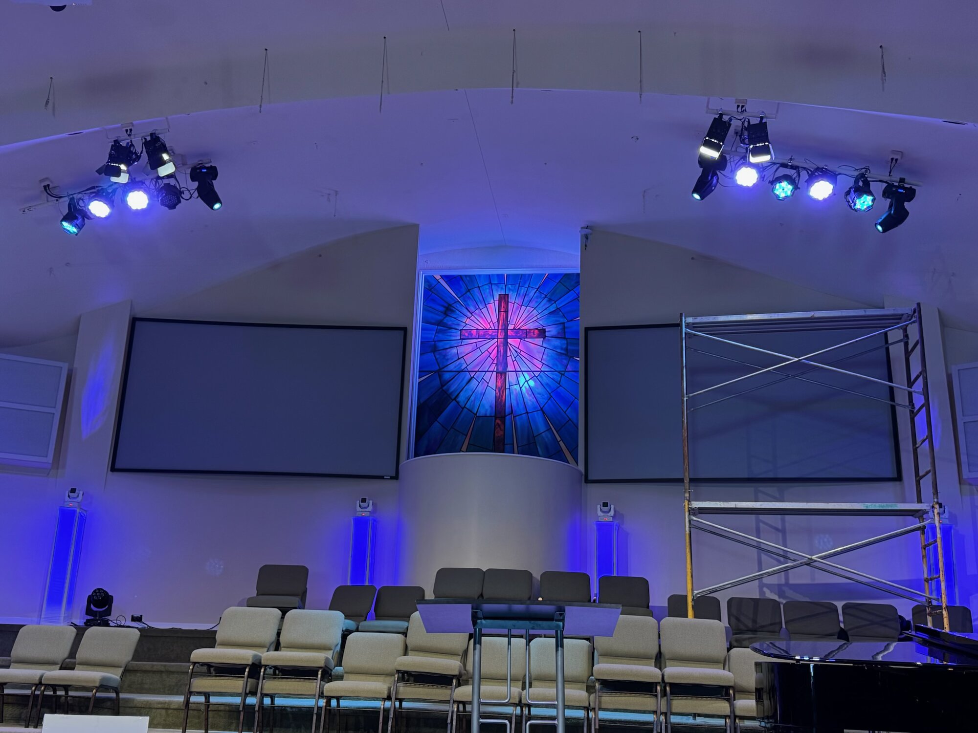 Install in progress at Parkview Baptist Church — a technician on a scaffold platform mounting overhead lighting fixtures with a deep blue light wash, stained-glass cross visible to the left.