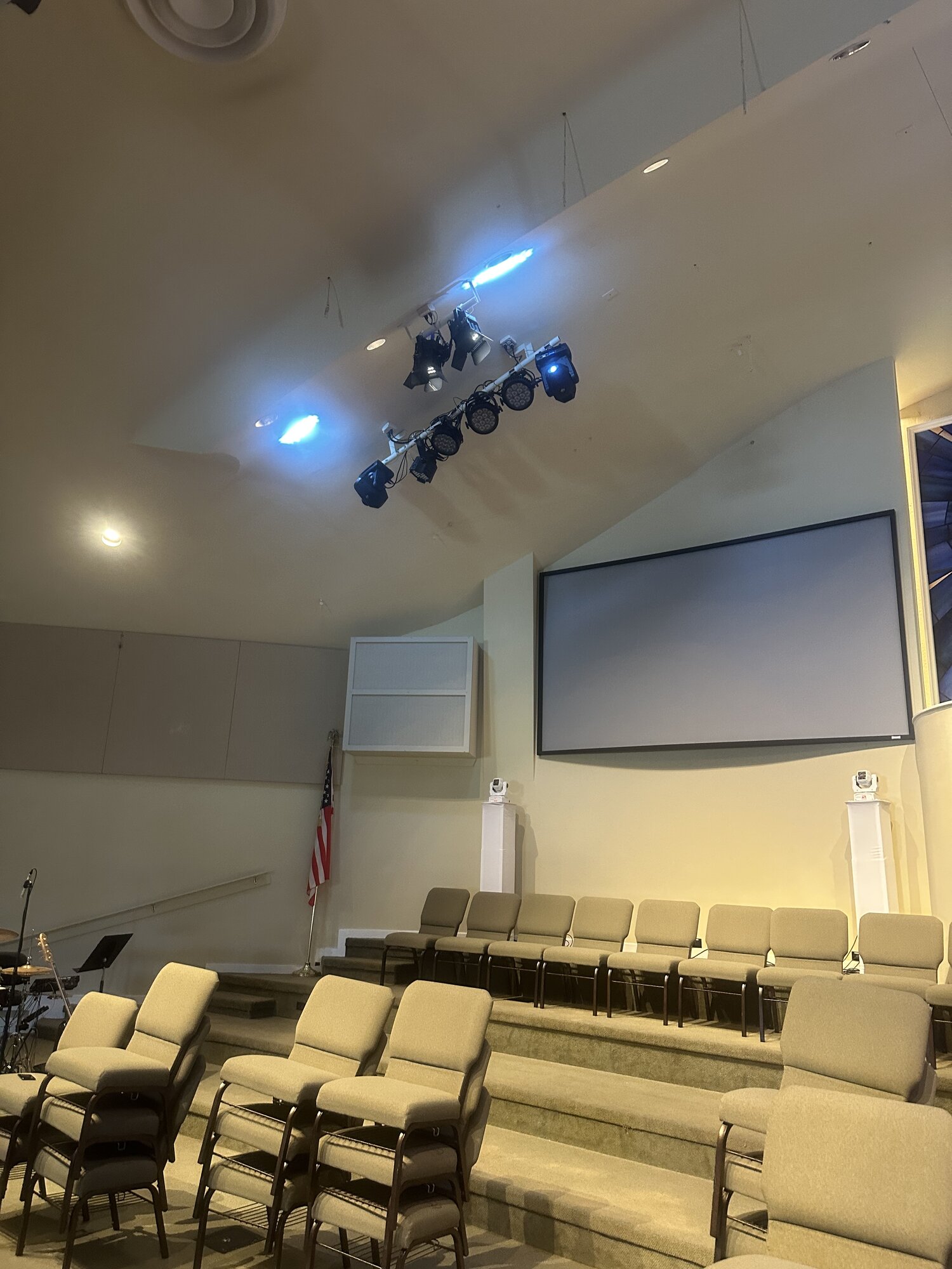 Parkview Baptist sanctuary in a coral-and-warm programmed state — vertical uplight columns washed in red-coral, the stained-glass cross at the back lit in matching tones, Elation PAR FC packaging visible on the platform from a recent install day.